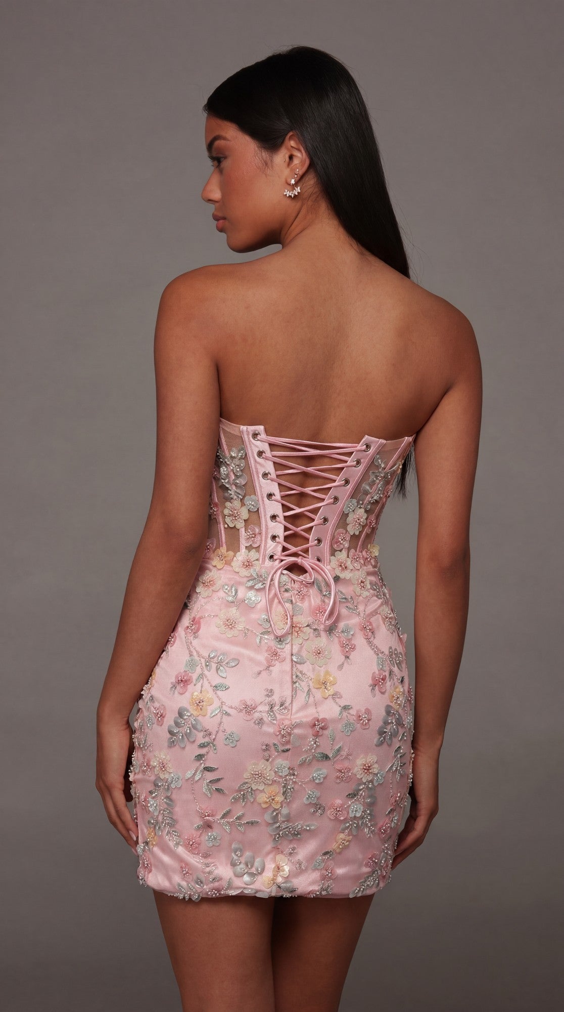Wearing the Alyce 40139 short homecoming dress, a woman poses against a plain gray background. The strapless pink dress features delicate floral appliques and a lace-up corset bodice.